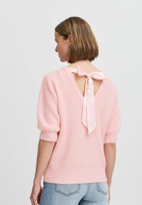 Roze gebreid sweater met een gestructureerde afwerking, korte mouwen en een V-hals aan de achterkant met een satijnen strikdetail. Gecombineerd met lichtblauwe jeans.