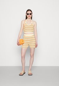 Conjunto de malha em croché de duas peças em padrão zigzag amarelo e laranja. Inclui um top curto e calções de cintura alta. Acompanhado de óculos de sol redondos e uma pequena mala laranja.