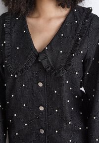 Camicia in tessuto trapuntato nero con colletto rifinito da ruches, decorata con piccole perle bianche e bottoni d'argento lungo la parte anteriore.