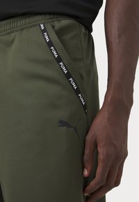 Olivengrønne træningsbukser med et sort Puma-logo, elastisk talje og en sort-hvid mærkestribe på siden.