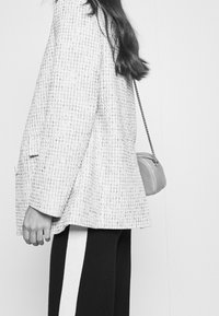 Šedý tvídový blejzr s volným střihem a texturovaným vzorem, doplněný černými kalhotami s bílými bočními pruhy a malou šedou kabelkou.