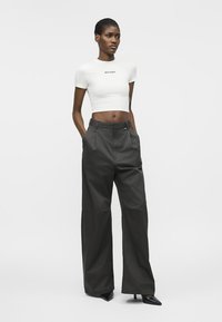 Γυναίκα που στέκεται με τα χέρια στις τσέπες, φορώντας λευκό κοντομάνικο τοπ και μαύρο παντελόνι ψηλόμεσο, με φαρδιά πόδια, και μαύρες μυτερές γόβες.