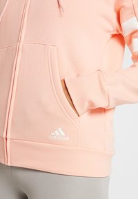 Felpa Adidas rosa chiaro con zip frontale, tasca a marsupio, polsini a coste e tre strisce bianche sulla manica. Presenta il logo sull'orlo.