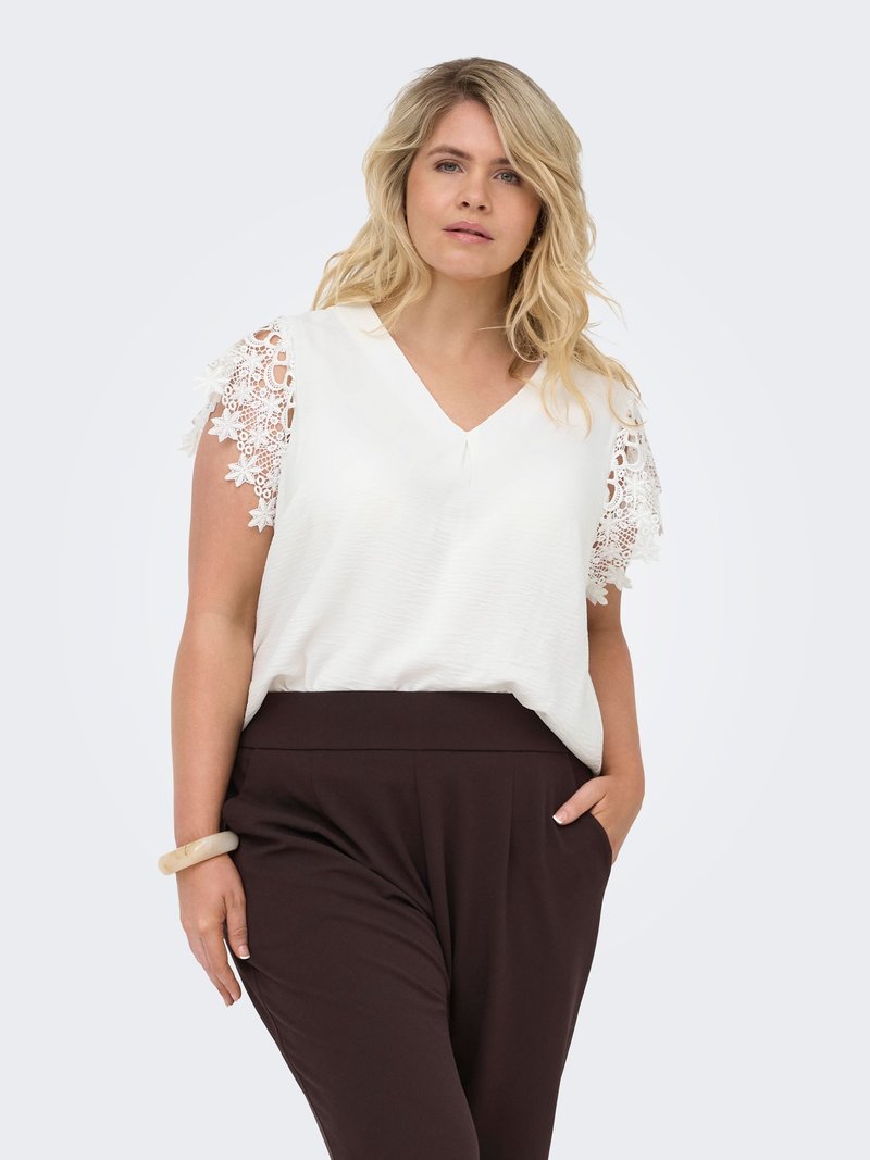 Femme aux cheveux blonds portant un chemisier blanc à manches en dentelle et un pantalon taille haute foncé, posant avec une main dans la poche et une expression neutre.