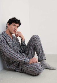 Conjunto de pijama a cuadros en tonos azules, rojos y blancos, hecho de una tela ligera. Un hombre está sentado contra una pared, con calcetines grises texturizados en los pies.