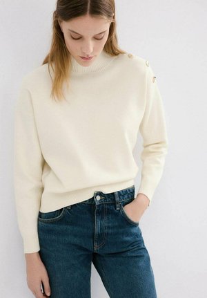 Cremefarbener Strickpullover mit hohem Kragen und gerippten Bündchen, mit goldenen Knöpfen auf der Schulter, kombiniert mit dunkelblauen Denim-Jeans.