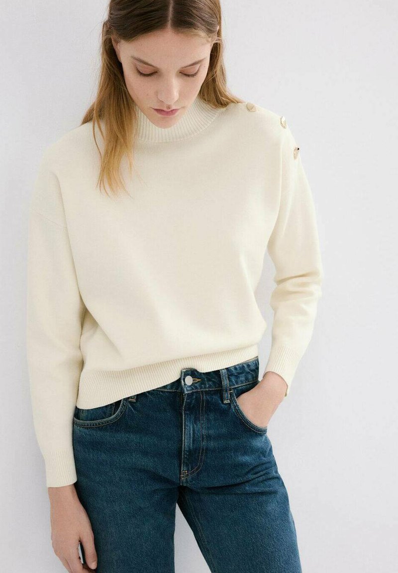 Pulover crem tricotat cu guler înalt și manșete cu dungi, având nasturi aurii pe umăr, purtat cu blugi denim albastru închis.