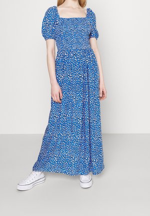 Une femme portant une robe maxi bleue à motifs floraux avec des manches bouffantes et des baskets blanches, debout devant un fond blanc uni.