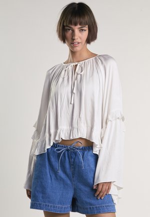 Junge Frau trägt eine lockere weiße Bluse mit Rüschen und blaue Jeansshorts, die an der Taille gebunden sind, und steht vor einem einfarbigen Hintergrund.