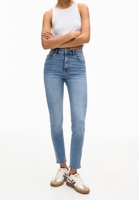 Vaqueros de talle alto de color azul claro con corte ajustado, con un dobladillo sin terminar. Combinados con una blusa sin mangas blanca y zapatillas blancas con detalles en marrón.