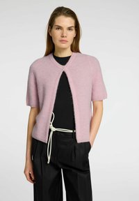 Lichtroze cardigan met korte mouwen, v-hals, open voorkant en een zachte, harige textuur. Draagt over een zwart shirt met een witte koordtaille.