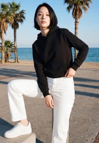 Donna con capelli corti neri, felpa nera e pantaloni bianchi che posa all'aperto vicino a palme sulla spiaggia in una giornata di sole.