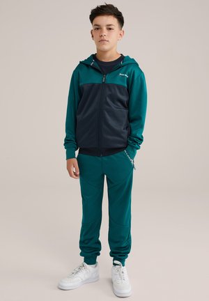 Teal och marinblå träningsoverall med blixtlåsjacka, elastiska muddar och matchande joggers. Har vita sneakers accentuerade och subtila designlinjer.