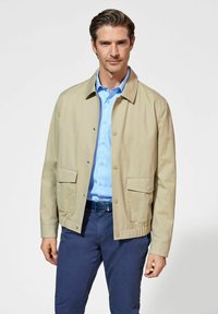 Beżowa, lekka kurtka z dwoma przednimi kieszeniami, zapi ęciem na zatrzask i elastycznym dołem, noszona na jasnoniebieskiej koszuli i granatowych spodniach.