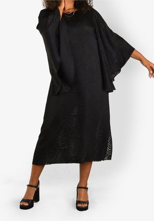 Vrouw die een zwarte, gestructureerde midi-jurk draagt met brede, vloeiende mouwen en zwarte plateauhakken, staande met één uitgestrekte arm.