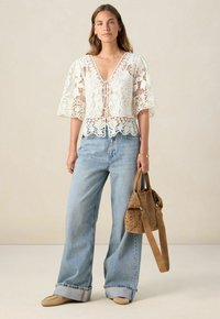 Weißes Spitzentop mit Glockenärmeln, tiefem V-Ausschnitt und gewelltem Saum, kombiniert mit hellblauen weit geschnittenen Jeans und einer braunen Handtasche mit Nieten.