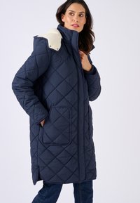 Marineblauwe gewatteerde lange jas met een hoge kraag, crèmekleurige accenten op de capuchon en zijzakken. Zachte textuur met een gestroomlijnd ontwerp.