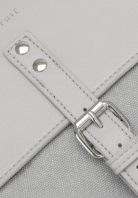 Harmaa nahkavyö, jossa on hopeiset kiinnitykset, niteille ja ompelulle, kiinnitettyä teksturoidun vaaleanharmaan kankaan taustalle.