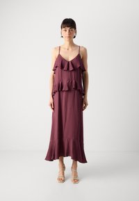 Langes, ärmelloses Kleid in Burgunderrot mit geschichteten Rüschen an der Vorderseite. Glatte Textur, fließendes Design und ausgestellter Saum. Getragen mit beigen Sandalen mit Riemchen.