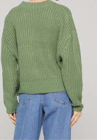 Personne portant un pull en maille épaisse vert clair et un jean en denim bleu, vue de dos sur un fond uni.