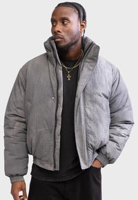 Gray padded jacket with a high collar, zip closure, and snap buttons. Features ribbed cuffs and a textured surface. Worn over a black shirt.