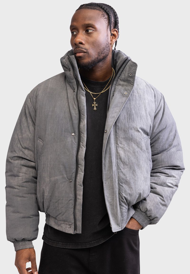 Gray padded jacket with a high collar, zip closure, and snap buttons. Features ribbed cuffs and a textured surface. Worn over a black shirt.
