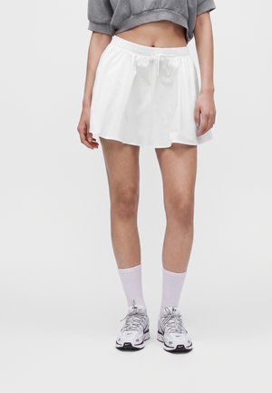Femme portant une jupe de sport blanche, un sweat court gris, des chaussettes blanches et des baskets blanches Nike, debout devant un fond uni.