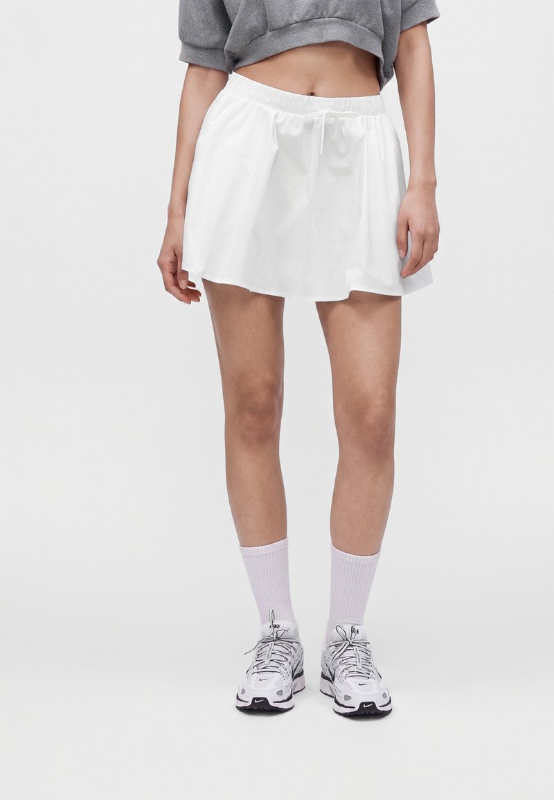 Femme portant une jupe de sport blanche, un sweat court gris, des chaussettes blanches et des baskets blanches Nike, debout devant un fond uni.