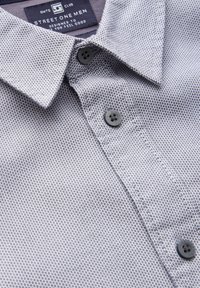 Camicetta grigia a maniche corte con un motivo geometrico piccolo e testurizzato, colletto alla francese e bottoni in plastica grigio scuro. Etichetta del marchio visibile.