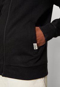 Hoodie noir zippé en coton, doté d'une poche kangourou, de poignets et d'un ourlet côtelés. Comprend une petite étiquette avec le texte "REBEL".