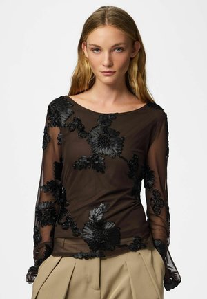 Femme aux longs cheveux blonds portant un top noir transparent avec une grande broderie florale et un pantalon beige plissé, debout sur fond blanc.