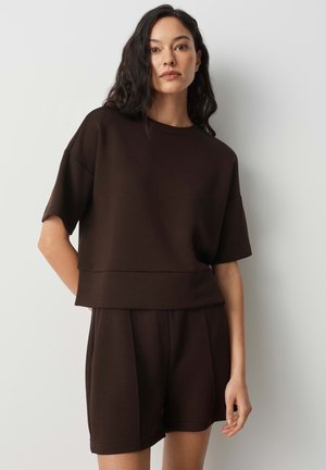 Woman wearing a dark brown short-sleeve top and matching shorts, standing against a plain light background with one hand behind her back.