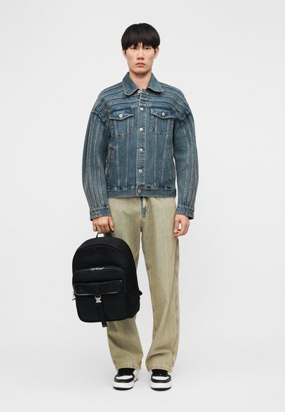 Νεαρός άνδρας που φοράει ένα μπλε ριγέ τζιν μπουφάν και μπεζ τζιν παντελόνι με φαρδιά μπατζάκια, κρατώντας μια μαύρη τσάντα πλάτης Off-White στο αριστερό του χέρι.