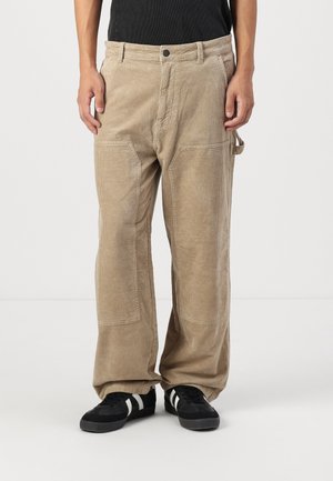 Pantalons en velours côtelé kaki avec une coupe décontractée, dotés de poches avant, d'une boucle latérale et d'un tissu ribbed texturé. Baskets noires avec des rayures blanches.