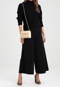 Donna che indossa un maglione nero a collo alto, pantaloni neri a gamba larga, tacchi con stampa leopardata e porta una borsa a tracolla beige con catena.