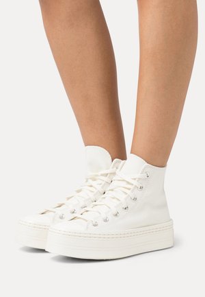 Baskets montantes blanches à plateforme avec semelles épaisses portées sur des jambes nues, vues de côté sur un fond uni.