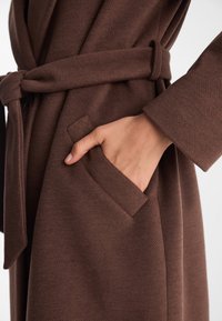 Brązowy płaszcz z wełny z teksturowanym wykończeniem, z paskiem w talii i bocznymi kieszeniami; ma długie rękawy i luźny krój.