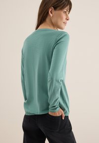 Chemise à manches longues en teal, fabriquée dans un tissu doux. Le dos présente un col rond et une coupe décontractée, associée à un pantalon noir.