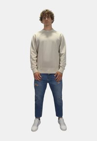 Sudadera de manga larga beige, con puños y dobladillo de canalé, combinada con jeans azules desgastados. El modelo lleva zapatillas blancas y está de pie sobre un fondo sencillo.