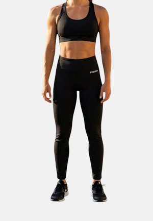 Persona in forma che indossa un reggiseno sportivo nero, leggings a vita alta con logo "FREDDY" e scarpe sportive nere, in piedi con le braccia rilassate lungo i fianchi.