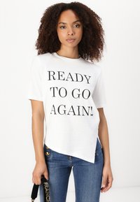 Balta medvilnė T-shirt su tekstu "PASIRUOŠĘS VĖL!" juoda spalva, trumpomis rankovėmis ir šiek tiek asimetrine apačia. Dėvima su mėlynomis džinsomis.