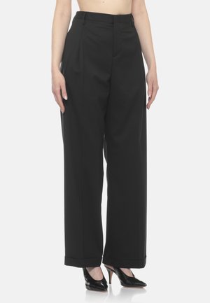 Donna che indossa pantaloni neri a vita alta, a gamba larga, con risvolti e tacchi neri con punta appuntita su uno sfondo chiaro e uniforme.