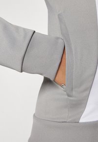Gray sports jacket featuring ribbed cuffs, zippered pocket, and contrasting white panel. Smooth texture with a fitted design.