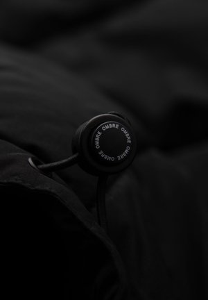 Sort stof med en tekstureret overflade, der har en rund lukning med toggle. Toglen har "OMBRE" trykt i hvidt omkring kanten.