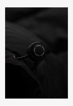 Sort stof med en tekstureret overflade, der har en rund lukning med toggle. Toglen har "OMBRE" trykt i hvidt omkring kanten.