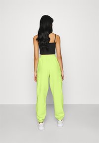 Kvinna med långt svart hår iklädd en svart crop top, neongröna sweatpants och vita sneakers, stående vänd mot en enkel vit vägg.