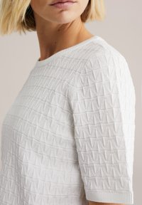 Biały teksturowany sweter z geometrycznym wzorem, okrągłym dekoltem i krótkimi rękawami. Tkanina wydaje się miękka i lekko wypukła.