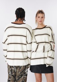 Ριγέ υπερμεγέθη πουλόβερ σε εκρού με καστανές οριζόντιες γραμμές, με ραβδωτή υφή, φαρδιά μανίκια και στρογγυλή λαιμόκοψη, σε συνδυασμό με διάφορα παντελόνια.