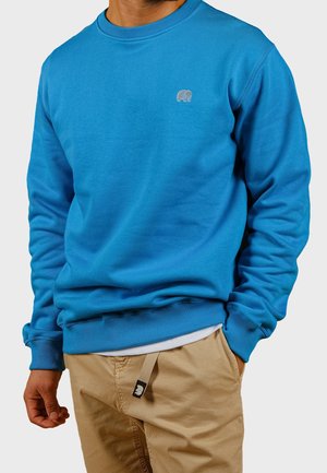 Mann trägt ein hellblaues Sweatshirt mit Rundhalsausschnitt und kleinem grauem Elefantenlogo auf der Brust sowie beige Hosen mit beigem Gürtel.