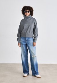 Grauer Pullover mit Dalmatinerkragen und gesticktem Logo, kombiniert mit hellblauen, weit geschnittenen Jeans mit Destroyed-Details, weißen Sneakers und Sonnenbrille.